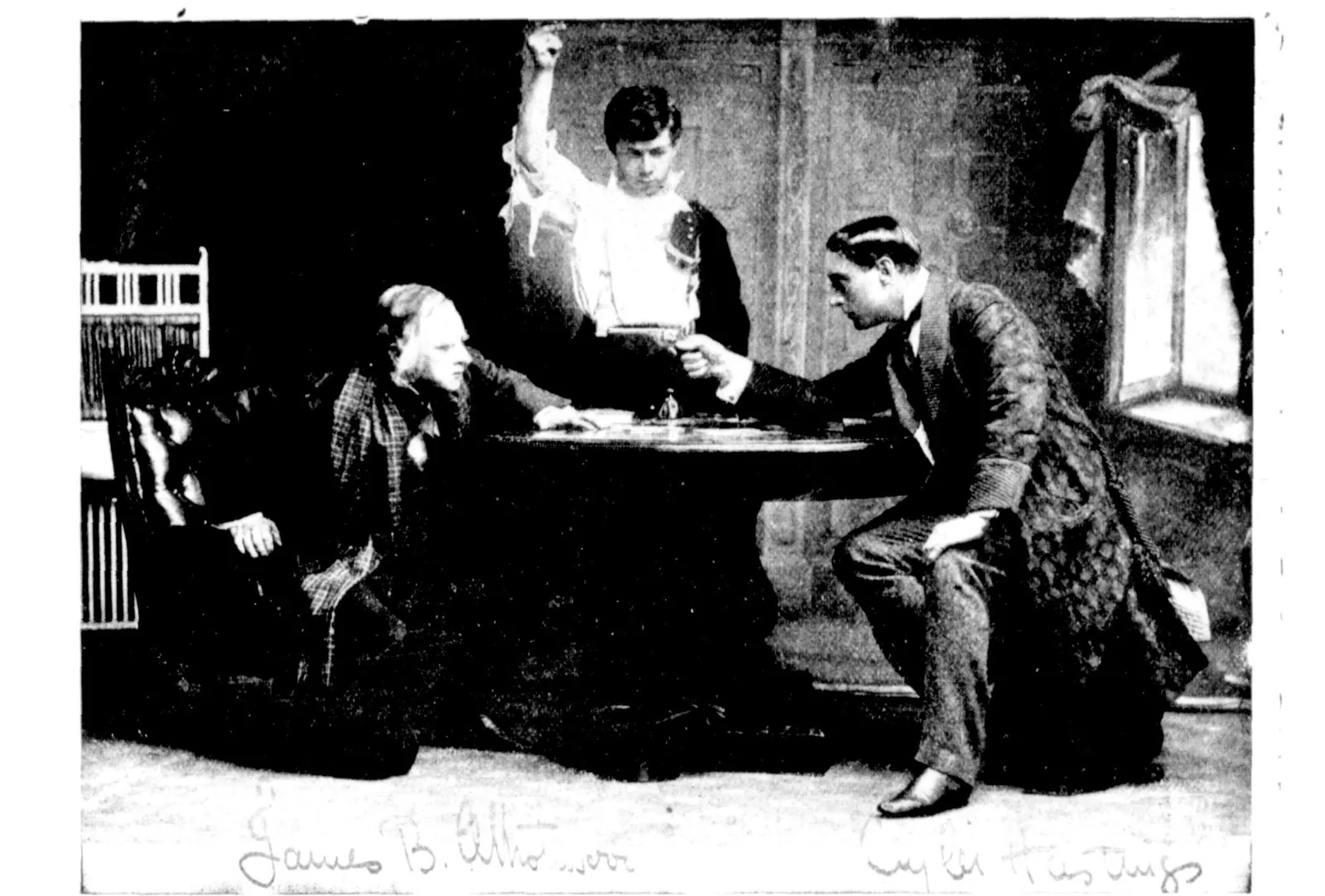 A stage photograph: Moriarty, seated at a table in a plaid shawl, faces Holmes, who leans across the table towards him, while Billy, behind them, raises his arm. Signed across the foot of the picture “James B. Atholwood” and “Cuyler Hastings”.